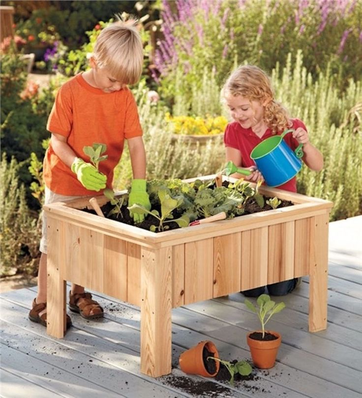 Two children garden at a wooden raised planter; one shovels soil while the other waters plants.